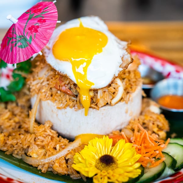 Thai fried rice served in a coconut shell, topped with a fried egg and a pink umbrella.