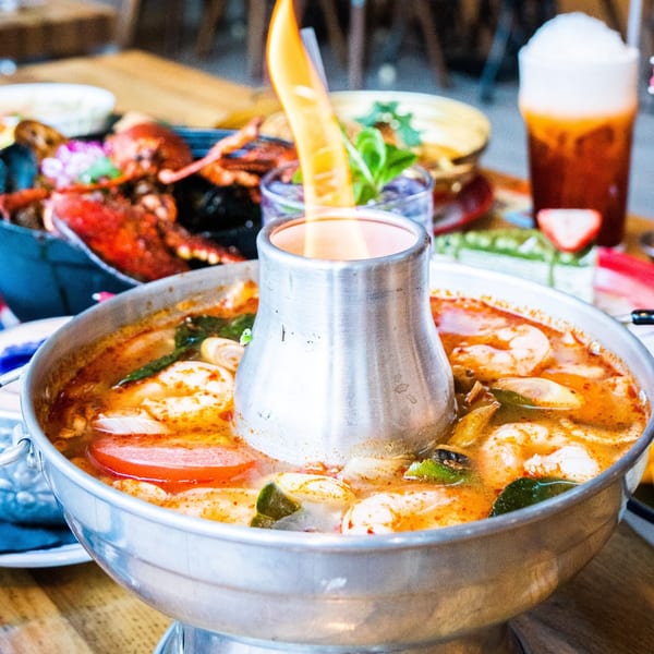A flaming hot pot of Tom Yum soup with shrimp and vegetables, alongside other Thai dishes.