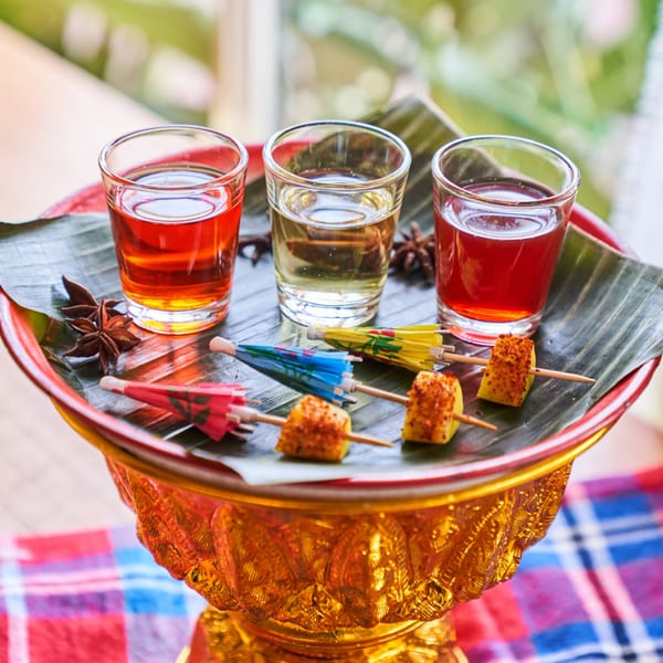 Three colorful Thai shots and pineapple skewers with mini umbrellas on a golden tray.