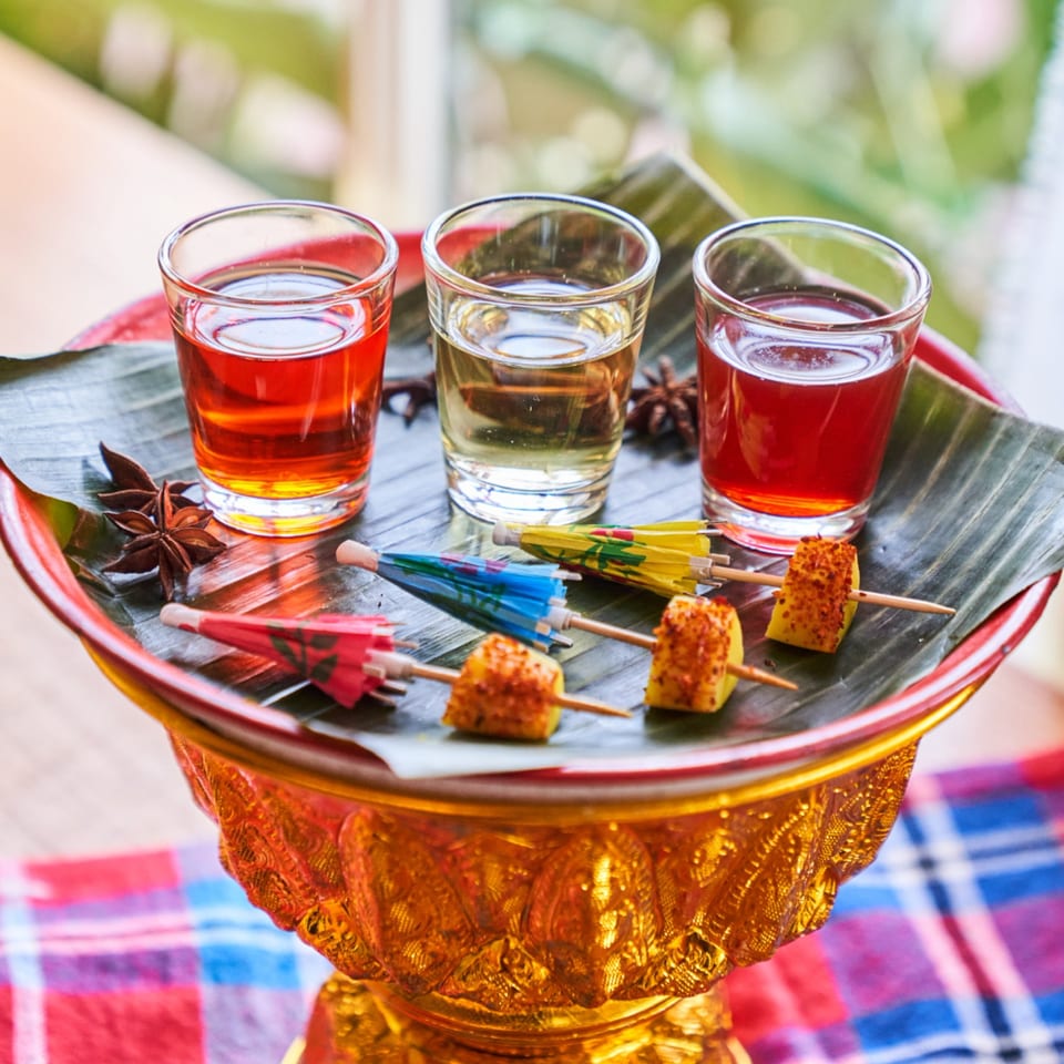 Three colorful Thai shots and pineapple skewers with mini umbrellas on a golden tray.